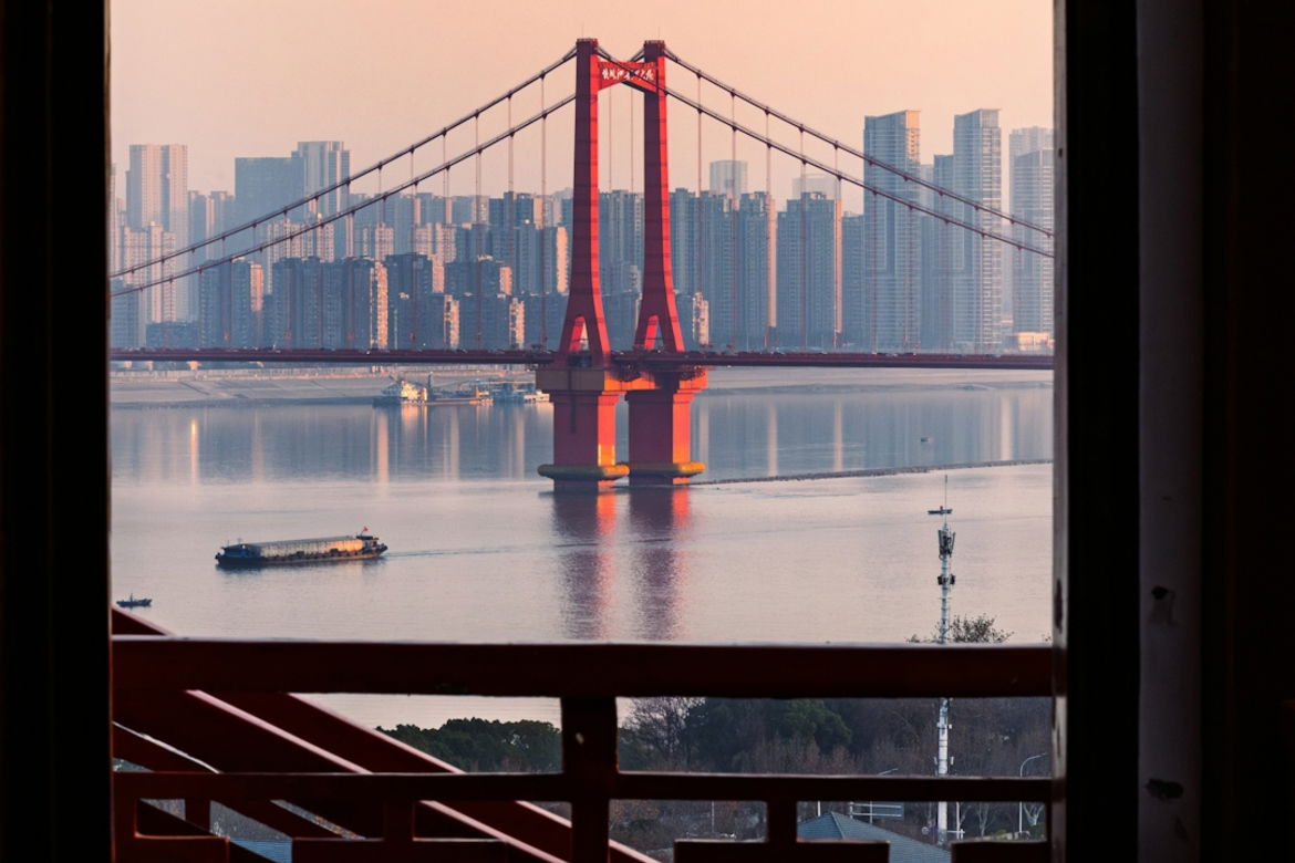 red-suspension-bridge-over-a-river-with-city-skyline-.jpg Red suspension bridge over a river with city skyline.